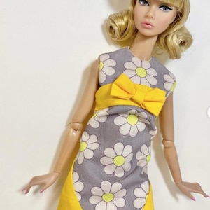 May include: A doll wearing a gray dress with white daisy patterns and a yellow bow. The dress has a yellow trim. The doll has blonde hair and heart-shaped sunglasses. The doll is wearing white tights.