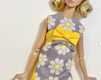May include: A fashion doll with blonde hair styled in a bob, wearing white heart-shaped sunglasses on its head. The doll is dressed in a sleeveless grey shift dress featuring a white daisy pattern with yellow centers, a yellow waist bow, and yellow side panels.
