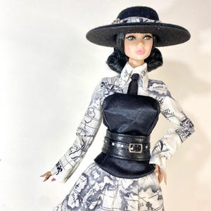 Könnte beinhalten: Eine Modepuppe mit einem schwarzen Hut mit einem schwarz-weiß gemusterten Band. Die Puppe trägt ein schwarz-weißes Outfit mit Kartenmuster, ein schwarzes Korsett und einen schwarzen Gürtel mit silbernen Akzenten.