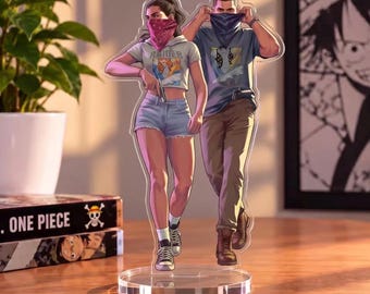 May include: A clear acrylic standee featuring two illustrated figures walking forward. The left figure wears a light-colored cropped hoodie, denim shorts, and sneakers, with a pink bandana. The right figure wears a blue t-shirt, brown pants, and boots, with a dark bandana.