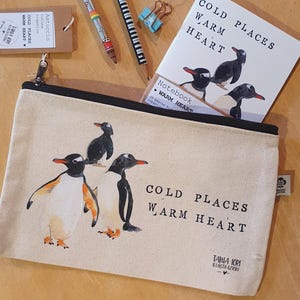 Può includere: Astuccio in tela beige con illustrazione di pinguini e la scritta "COLD PLACES WARM HEART". Include anche un taccuino, matite e una graffetta.
