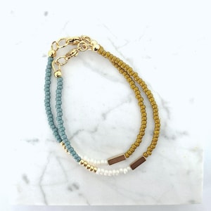 Op de afbeelding: Twee armbanden met kralen en goudkleurige sluitingen. Eén armband is lichtblauw en de andere mosterdgeel. Beide armbanden hebben goudkleurige accenten en zijn gemaakt van kleine, ronde kralen. De armbanden worden gepresenteerd op een wit marmeren oppervlak.