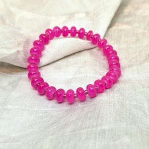 Può includere: Un braccialetto di perline rosa acceso. Il braccialetto è composto da perline rotonde e lucide, infilate insieme per formare un anello continuo. Le perline sono di un rosa brillante e saturo, e il braccialetto è appoggiato su un tessuto chiaro.