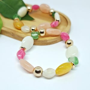 Puede incluir: Dos pulseras de cuentas con cuentas coloridas de forma irregular en rosa, blanco, amarillo y verde. Las pulseras tienen detalles metálicos dorados. Las pulseras se exhiben en una bandeja de madera.