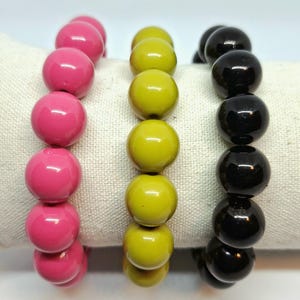 May include: Three beaded bracelets in vibrant colors: hot pink, olive green, and black. Each bracelet features large, round beads strung together. The bracelets are displayed on a white, textured surface.