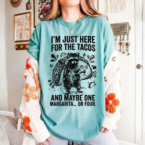 May include: A light blue t-shirt with the text "I'M JUST HERE FOR THE TACOS AND MAYBE ONE MARGARITA... OR FOUR." A black and white graphic of a raccoon holding a margarita and a taco is printed on the front.