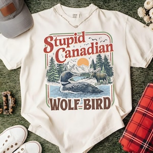 May include: Cream-colored t-shirt with a graphic that reads "Stupid Canadian Wolf Bird." The graphic features a loon on water, mountains, trees, and a moose. Other items include a gray cap, white sneakers, and a red plaid blanket.