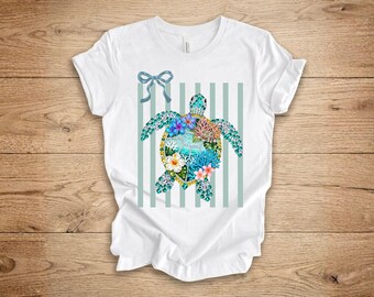 May include: White t-shirt featuring a sea turtle design. The turtle is decorated with colorful flowers and coral, set against a backdrop of vertical stripes and a blue bow. The design incorporates shades of blue, pink, and green.