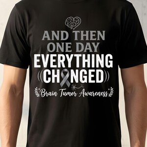 Może przedstawiać: Czarny t-shirt z białym i szarym napisem "AND THEN ONE DAY EVERYTHING CHANGED". Zawiera szarą wstążkę i grafikę mózgu oraz tekst "Brain Tumor Awareness".