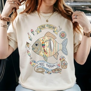 Può includere: T-shirt color crema con un design di pesce colorato con le scritte "GONE FISHIN!" e "REEL THERAPY." Il pesce è circondato da esche da pesca e elementi floreali. La maglietta è realizzata in un materiale morbido.