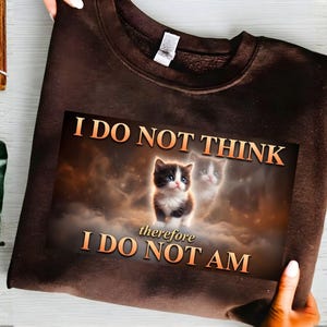 May include: A brown sweatshirt featuring a graphic of a kitten in a cloudy background with the text "I DO NOT THINK therefore I DO NOT AM" in orange letters. The sweatshirt is held up by a person.