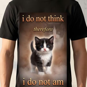 May include: Black t-shirt featuring a kitten with black and white fur and blue eyes. The shirt has the text "i do not think therefore i do not am" in orange and white. The background has a blurred image of a cat.