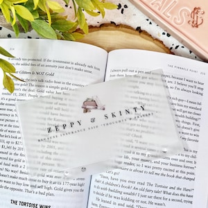 May include: A rectangular, semi-transparent frosted bookmark featuring the text ZEPPY & SKINTY and the phrase BECAUSE INSURANCE SAID THOUGHTS & PRAYERS. The bookmark includes a small graphic of a pen and a notebook, resting on an open book page.