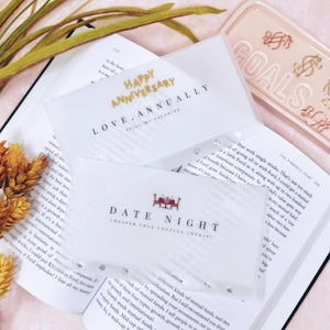 May include: Clear plastic cards with gold text reading "HAPPY ANNIVERSARY" and "DATE NIGHT" are displayed on an open book. A pink tray with the word "GOALS" and dollar sign charms is in the background. Dried flowers add a decorative touch.