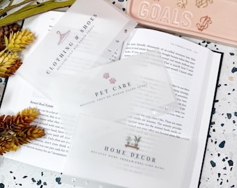 May include: Three translucent plastic cash envelopes labelled CLOTHING & SHOES, PET CARE, and HOME DECOR with corresponding icons, resting on an open book next to a pale pink rectangular tray with the word GOALS and four gold dollar sign clips.