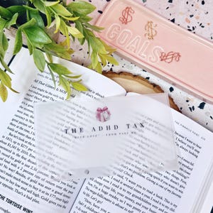 May include: A translucent, frosted rectangular card featuring a pink gift icon and the black text "THE ADHD TAX" followed by "WITH LOVE - FROM PAST ME". The card is positioned over an open book, near a pink ceramic tray with the word "GOALS" embossed.