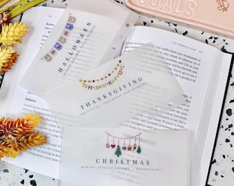 May include: Transparent plastic inserts featuring festive text and illustrations. The inserts display "Halloween," "Thanksgiving," and "Christmas" designs. They are positioned over an open book, with a pink container marked "GOALS" visible.