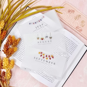 May include: Clear plastic planner inserts labeled July, August, and September, with decorative graphics. The inserts are on top of an open book, with a pink tray that says "GOALS" in the background.