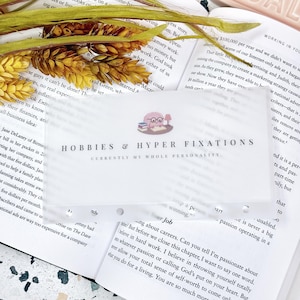 May include: A translucent rectangular planner divider featuring a cartoon brain character wearing glasses at a desk with books. The text reads HOBBIES & HYPER FIXATIONS and CURRENTLY MY WHOLE PERSONALITY. The divider has punched holes along the bottom edge.