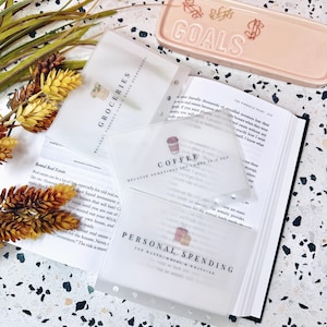 May include: Three translucent plastic cash envelopes for a binder, labeled Groceries, Coffee, and Personal Spending with small icons. Each envelope features black text and a graphic. A peach-colored tray with the word Goals is visible nearby.