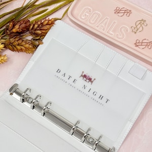May include: White binder with a clear pocket displaying "DATE NIGHT" text and a small graphic of a table setting. The binder has a silver metal ring mechanism. A pink tray with the word "GOALS" is in the background.