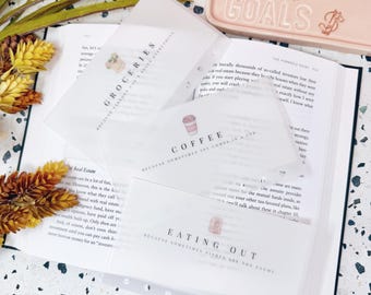 May include: Three translucent white plastic cash envelopes with black text and small icons. The envelopes are labelled GROCERIES, COFFEE, and EATING OUT, each with a corresponding small illustration of a grocery bag, a coffee cup, and a takeout box.