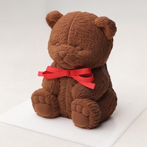 May include: A brown teddy bear with a red ribbon tied in a bow around its neck. The bear has a textured, velvety appearance and is sitting on a white surface. The bear's features include small ears, a rounded snout, and paws.