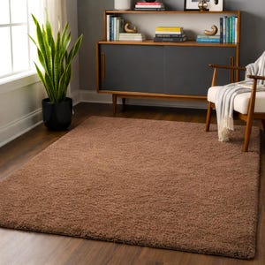 Puede incluir: Una alfombra rectangular de pelo largo marrón. La alfombra está colocada sobre un suelo de madera. Una planta en maceta negra está en la esquina de la habitación. Un mueble y una silla de madera están en el fondo.