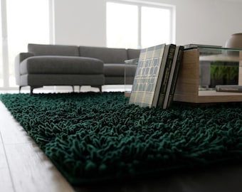 May include: A deep forest green shag area rug with a thick, textured pile. The rug features long, soft fibers that create a dense, plush surface covering the floor. The rug is positioned under a wooden coffee table and a gray sofa.