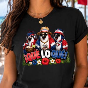 Peut inclure: Un t-shirt noir avec un motif représentant trois perroquets portant des lunettes de soleil et des chapeaux, accompagné du texte "¡QUÉ LO QUÉ!". Les perroquets sont ornés de rouge, blanc et bleu, avec des fleurs et des feuilles tropicales.