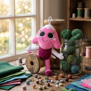 K&ouml;nnte beinhalten: Ein Pl&uuml;schtier in Form eines rosa Berges mit wei&szlig;er Spitze, gro&szlig;en schwarzen Augen und einem l&auml;chelnden Mund. Das Spielzeug hat wei&szlig;e Arme und dunkelrosa Beine und sitzt auf einer Holzspule. Daneben liegen gr&uuml;ne Wollkn&auml;uel in einem Glas.