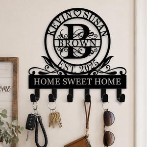 May include: A black metal wall-mounted key holder featuring a circular monogram with the letter D, the names Kevin and Susan, and the text Est. 2025. Below the circle, a rectangular plaque displays the text Home Sweet Home with six hooks.