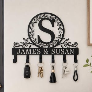 Könnte beinhalten: Ein schwarzer Wand-Schlüsselhalter aus Metall mit einem kreisförmigen Rahmen, der in der Mitte einen großen Buchstaben S zeigt, umgeben von einem Blattmuster. Unter dem Kreis befindet sich eine Leiste mit dem Text JAMES & SUSAN und sechs Haken.