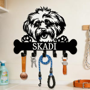 May include: Black metal key holder featuring a dog design with the name "SKADI". Includes hooks for keys and leashes. Accessories include a blue leash, keys, and a leather tag. A decorative and functional item.