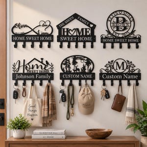 May include: Six black metal wall-mounted key holders with hooks. Designs include infinity symbols, house shapes, circular monograms, and nature scenes. Text includes "HOME SWEET HOME," "THE SMITH FAMILY," "KEVIN SUSAN BROWN," and "Johnson Family."