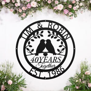 Könnte beinhalten: Schwarze Metall-Wandkunst mit den Namen "TIM & ROBIN" und dem Text "40 YEARS Together EST. 1986". Das Design zeigt zwei Vögel, die auf einem Ast über einem Herzen sitzen, alles in einem kreisförmigen Rahmen.