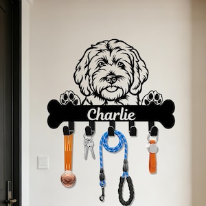Pode incluir: Um porta-chaves de metal preto com um design de cão e o nome "Charlie". O porta-chaves tem um design em forma de osso com ganchos para chaves e trelas. Vários itens penduram dos ganchos, incluindo chaves, uma trela e uma medalha.