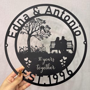 Può includere: Arte da parete in metallo nero con i nomi "Edna & Antonio" e il testo "30 anni insieme EST. 1996". L'arte presenta un albero con uccelli, una coppia su una panchina e forme di cuore, il tutto all'interno di una cornice circolare.