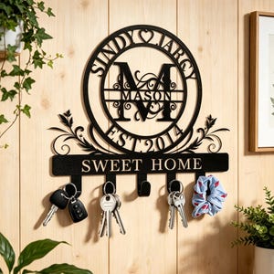 Könnte beinhalten: Schwarzer Metall-Schlüsselhalter mit dem Text "SWEET HOME" und Haken. Das Design umfasst ein kreisförmiges Monogramm mit dem Namen "MASON" und dem Jahr "EST 2014". Schlüssel und ein Scrunchie hängen an den Haken. Der Schlüsselhalter ist an einer Holzwand befestigt.