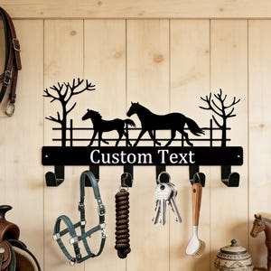 May include: Black metal key holder featuring a silhouette of two horses and the text "Custom Text". The key holder has five hooks and is decorated with trees and a fence. Various equestrian items hang from the hooks.