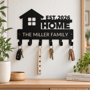 May include: A black metal wall-mounted key holder featuring a house silhouette, the text "EST. 2026", "HOME", and "THE MILLER FAMILY". It has six hooks, with four sets of keys hanging from them, including a beige fabric keychain with black hearts.