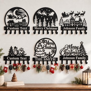 Könnte beinhalten: Sechs schwarze Wand-Schlüsselhalter aus Metall mit weihnachtlichen Motiven. Die Designs zeigen Silhouetten von Weihnachtsmann im Schlitten, Tannenbäumen, Schneemännern und Texten wie Custom Text, Merry Christmas und Johnson Family. Fünf Haken pro Leiste.