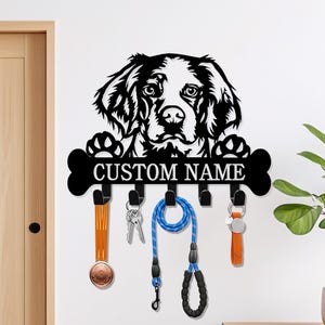 May include: Black metal dog-themed key holder with the text "CUSTOM NAME". The key holder features a dog's head and paws above a bone-shaped section with hooks. Hanging from the hooks are keys, a dog leash, and other accessories.