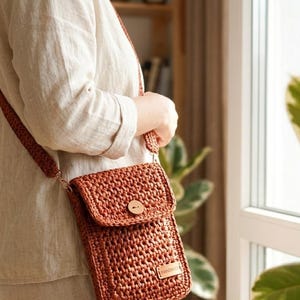 May include: A rust-coloured crochet crossbody bag featuring a rectangular shape, a fold-over flap secured with a single wooden button, and a matching crochet shoulder strap. A small rectangular label with the text "HANDMADE" is attached to the front.
