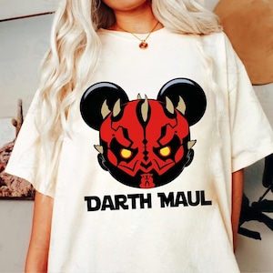 May include: Cream-colored t-shirt featuring a Darth Maul graphic. The graphic combines the character's likeness with Mickey Mouse ears, rendered in red, black, and yellow. The words "Darth Maul" are printed in bold, black letters below the graphic.