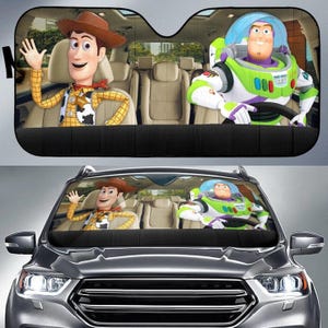 May include: Black car sunshade featuring Woody and Buzz Lightyear from Toy Story. Woody is wearing a cowboy hat and waving. Buzz is in his space suit. The sunshade is installed in a gray car.