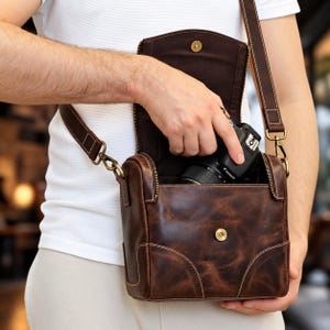Puede incluir: Una bolsa para cámara de cuero marrón oscuro con estructura rectangular, solapa con cierre de botón a presión color latón y correa ajustable con herrajes metálicos. La bolsa contiene una cámara digital negra con su objetivo.