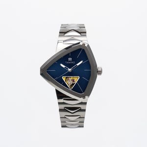 Op de afbeelding: Een roestvrijstalen horloge met een unieke driehoekige wijzerplaat en een diepblauwe wijzerplaat. Het horloge heeft de merknaam "TAMIRO" en een kleine, zichtbare mechanische beweging. De horlogeband is ook van roestvrij staal.
