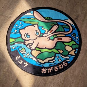 Peut inclure: Tapis rond avec une illustration de Mew de Pokémon. Le tapis a une bordure bleue avec du texte japonais et un motif de terre et d'eau. L'image centrale montre Mew en rose.