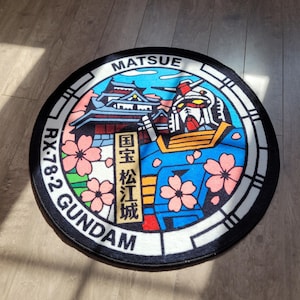 Könnte beinhalten: Ein runder Teppich mit einer grafischen Darstellung eines Gundam-Roboters und einer japanischen Burg. Das Design zeigt rosa Kirschblüten, einen blauen Himmel und den Text MATSUE, RX-78-2 GUNDAM sowie japanische Schriftzeichen. Schwarzer Rand.
